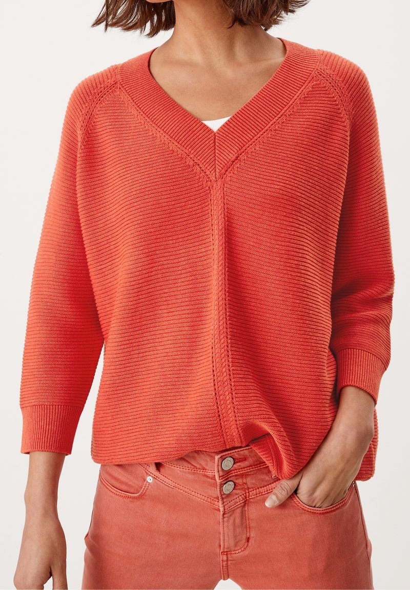 Pull léger de couleur orange vif avec un col en V et une texture côtelée. Il présente des manches trois-quarts et une coupe décontractée, associé à un jean rose.