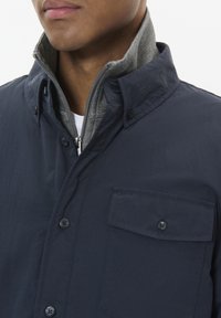 Chaqueta ligera azul marino con cuello alzado, con un forro interior gris, cierres de botones y un bolsillo en el pecho con solapa.