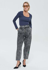 Femme portant des lunettes de soleil ambre, un haut à encolure large navy à manches longues, un jean taille haute en délavé et des escarpins noirs, se tenant avec les mains dans les poches.