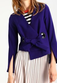 Fialová zavinovací bunda s detailem mašle a velkými černými kruhovými výřezy. Modelka má na sobě plisovanou krémovou sukni.