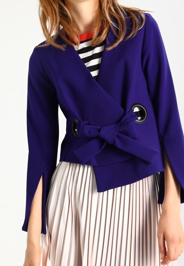 Fialová zavinovací bunda s detailem mašle a velkými černými kruhovými výřezy. Modelka má na sobě plisovanou krémovou sukni.