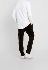 Person wearing a white long-sleeve shirt, dark brown corduroy pants, and white sneakers standing against a plain light background.