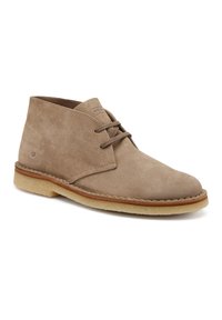 Stivale chukka in suede beige con punta rotonda, due fori per i lacci e una suola in gomma crema con un accento di cucitura lungo il bordo.