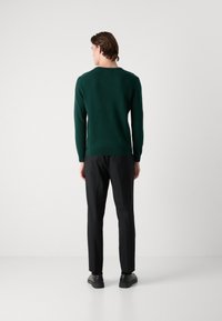 Puloverul verde închis are un guler rotund și manșete cu nervuri. Este purtat împreună cu pantaloni negri eleganți și pantofi negri, evidențiind un fit clasic și o textură netedă.