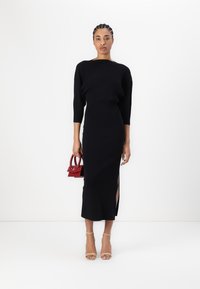 Un crop top noir à côtes associé à une robe noire longue et ajustée avec une fente sur le côté, complétée par un petit sac à main rouge et des talons beiges.