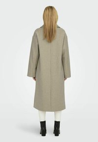 Lang grijs wollen coat met een rechte snit en brede mouwen, met een achterbeurt en een eenvoudig ontwerp. Zwarte enkellaarsjes zichtbaar eronder.