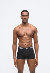 Μυώδης άνδρας όρθιος, στραμμένος προς τα εμπρός, φοράει μαύρα μποξεράκια Lacoste με λογότυπο στη μέση, σε απλό φωτεινό φόντο.