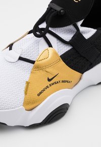 Nike sportssko med en hvid mesh-overdel, sorte accenter, en gylden hælpanel med teksten "GROOVE.SWEAT.REPEAT," og en tekstureret sort sål.