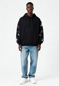 Felpa nera con cappuccio e tasca frontale, con vari accenti ricamati colorati. Abbinata a jeans larghi color blu chiaro e sneakers bianche.