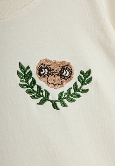 Ένας κρεμ κάλυμμα υφάσματος διατίθεται με λεπτομέρεια κεντημένη του προσώπου ενός χαρακτήρα περιτριγυρισμένου από πράσινα φύλλα. Ο σχεδιασμός περιλαμβάνει μεγάλα, εκφραστικά μάτια.