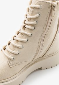 Bottine en cuir beige à lacets avec une fermeture éclair sur le côté. Elle possède une semelle robuste et une surface texturée avec des détails de couture.