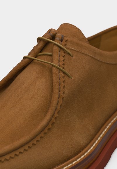 Κοντινό πλάνο καφέ δερμάτινου παπουτσιού από σουέντ με ραφή moc toe και στρογγυλά καφέ κορδόνια, που δείχνει λεπτομερώς την υφή και την άκρη της σόλας.