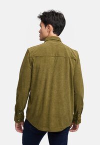 Camicia in velluto a coste verde oliva con maniche lunghe, bottoni sui polsini e dettaglio a yoke. Il tessuto testurizzato e la vestibilità su misura sono caratteristiche importanti.