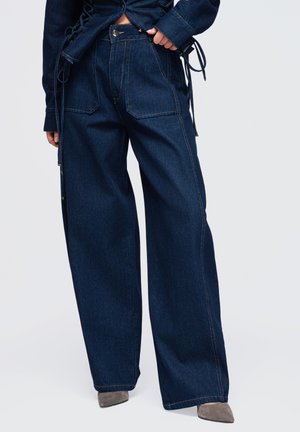 Personne portant un jean bleu foncé à jambes larges, assorti à une veste en jean assortie et des chaussures grises à bouts pointus, debout devant un fond blanc.