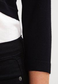 Zwarte gebreide trui met een opgerolde manchette, gedragen over een witte blouse. Donkere jeans zichtbaar bij de taille hebben een metalen knoop. Glad textuur.