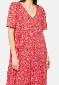 Femme portant une robe rouge avec de petits motifs abstraits blancs, à manches courtes et décolleté en V, visible de la moitié de la poitrine jusqu'en dessous de la taille.