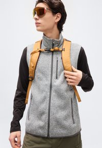 Νεαρός που φοράει αθλητικά γυαλιά ηλίου με ανακλαστικούς φακούς, γκρι γιλέκο fleece με φερμουάρ, μαύρα μακριά μανίκια, λουριά σακιδίου πλάτης μουσταρδί και χακί παντελόνι.