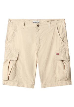 Uomo con pantaloncini cargo beige con tasche laterali, calzini bianchi e sneakers bianche, in piedi davanti a uno sfondo semplice.