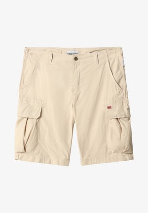 Homme portant un short cargo beige avec des poches latérales, des chaussettes blanches et des baskets blanches, debout devant un fond uni.