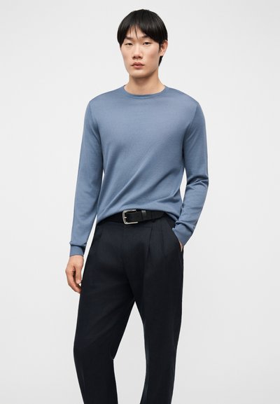 Νέος άνδρας που στέκεται με τα χέρια στις τσέπες, φοράει μπλε πουκάμισο με μακριά μανίκια και μαύρο παντελόνι με μαύρη ζώνη, μπροστά σε απλό φόντο.