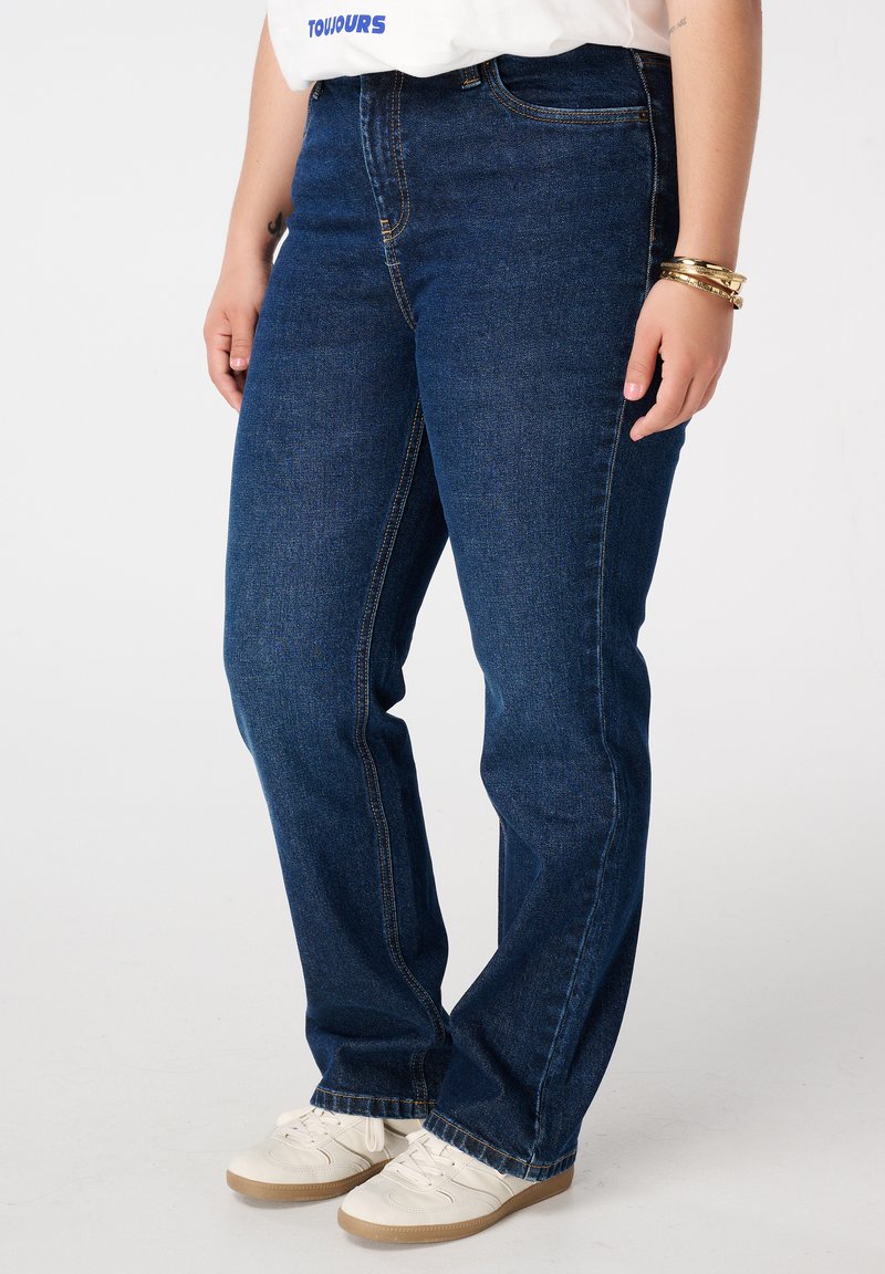 Donkerblauwe rechte spijkerbroek van denim, met een klassiek vijf-pocket ontwerp en zichtbare stiksels langs de naden. Draagt met witte sneakers.