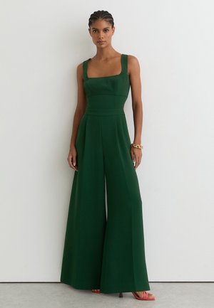 Femeie cu părul împletit purtând un salopetă verde închis fără mâneci și o brățară aurie, stând în fața unui perete alb simplu.