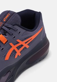 Vijolični športni čevelj z oranžnimi detajli, mrežasti zgornji del, gumijast podplat in značilna oblika logotipa na strani. Vidni teksturirani materiali.
