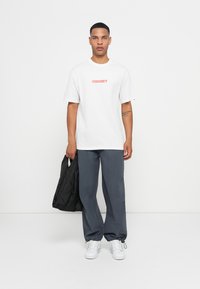 Young man stands holding a black tote bag, wearing a white Quiksilver T-shirt, gray pants with elastic cuffs, and white sneakers.