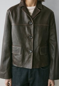 Brun læderjakke med krave, tre sorte knapper og to påsyede lommer. Glat tekstur med kontrasterende syningsdetaljer.