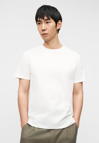 Jeune homme aux cheveux noirs portant un t-shirt blanc uni à manches courtes et un pantalon vert olive, debout les mains dans les poches devant un fond blanc.