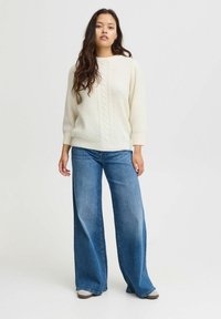 Pull en maille torsadée crème avec des manches trois-quarts associé à un jean bleu taille haute et large. Le mannequin se tient devant un fond neutre.