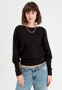 Un pull noir à coupe décontractée, avec des manches longues et un ourlet côtelé, associé à un jean clair taille haute. Un simple collier en chaîne en accent.