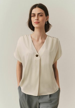 Žena s tmavými vlasmi po plecia nosí krémovú blúzku s výstrihom do V a jedným gombíkom, sivé nohavice, stojí s rukami v vreckách.