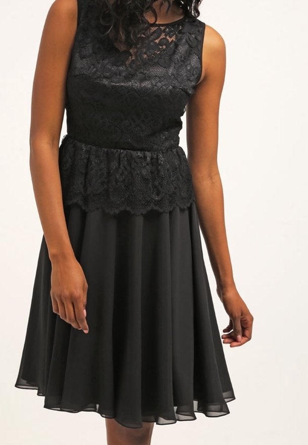 Svart klänning med spetsdetaljer på överkroppen, ärmlös design och peplum midja. Utrullad kjol av genomskinligt tyg med en vågig nederkant.