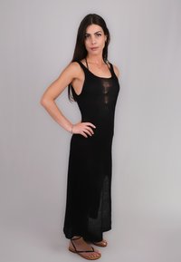 Abito maxi nero a maglia con design senza maniche, caratterizzato da un corpetto aderente e una texture trasparente. Indossato con sandali piatti marroni.