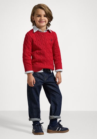 Roter Cable-Knit-Pullover, darunter ein blau-weißes Hemd mit Kragen, dunkle Jeans, an den Bündchen aufgerollt, und marineblaue Sneaker mit weißen Streifen.