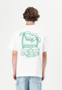 Bela bombažna majica s kratkimi rokavi, ki na hrbtni strani vsebuje zeleno grafiko palm, valov in besedila "adidas Originals". Ima sproščen kroj.