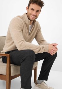 Beiger Pullover mit halbem Reißverschluss aus weichem Stoff, kombiniert mit dunkelgrauer Hose. Das Modell sitzt auf einem Stuhl und trägt beige Sneaker und graue Socken.