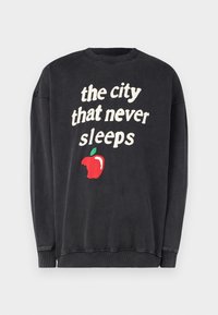 Sudadera negra de manga larga, cuello redondo y con el texto "la ciudad que nunca duerme" acompañado de un gráfico de una manzana roja. Material de algodón, textura suave.