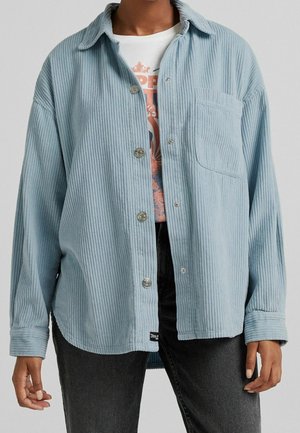 Osoba ubrana w jasno-niebieską prążkowaną kurtkę zapinaną na guziki, na koszulce z nadrukiem i czarnych dżinsach, stojąca z rękami swobodnie opuszczonymi wzdłuż ciała.