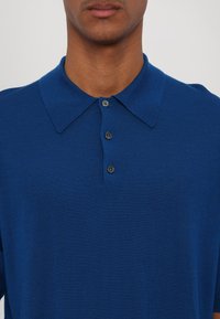 Niebieska koszulka polo wykonana z teksturowanej tkaniny, z klasycznym kołnierzykiem i trzema guzikami w ciemnym kolorze.