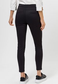 Pantaloni skinny neri con una vita alta, caratterizzati da due tasche posteriori, abbinati a sneakers nere su uno sfondo chiaro.