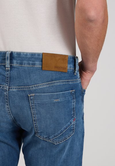 Person wearing blue denim jeans with a back pocket and brown leather brand patch on the waistband, hand resting in front pocket.