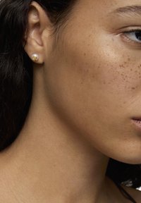 Kolczyki na sztyftach w złotym odcieniu mają perłowo-białą przednią część oraz unikalny teksturowany akcent. Włosy są ciemne, falowane i częściowo schowane za uchem.