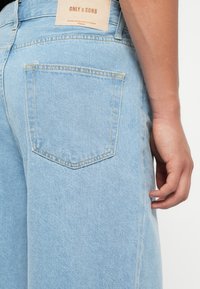 Lys blå denimjeans med vanlig passform, med en baklomme og et merket lærpatch over. Teksturen er glatt med synlig søm.