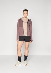 Lett mauve hettejakke med to frontlommer over en beige t-skjorte. Svart shorts og grå sneakers med hvite lisser og gummisåle.
