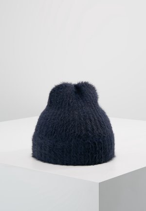 Tummansininen, karhea neulottu pipo, jossa on käännetty resori, esillä valkoisella jalustalla yksinkertaisen vaaleanharmaan taustan edessä.