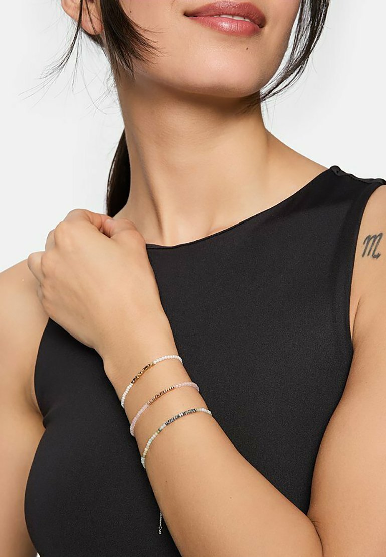 Armband med en kombination av små kulor i vitt, rosa och metalliska toner av guld och silver, bärs på ett handled. En svart ärmlös topp synlig.