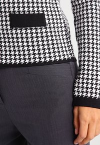 Svart och vit houndstooth-mönstrad stickad jacka med en svart ficka och en figurnära design, kombinerad med mörkgrå skräddarsydda byxor.