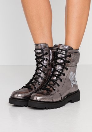 Błyszczące metaliczne botki sznurowane na kostkę z czarnymi podeszwami, czarnymi sznurowadłami i logo "KARL" na języku i boku, noszone na nagich nogach.
